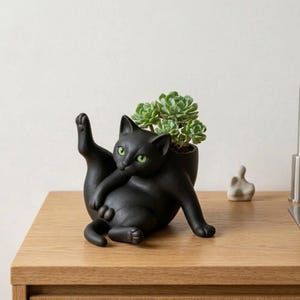 Op de afbeelding: Een zwarte katvormige plantenbak met groene ogen, die zijn poot omhoog houdt, en een vetplantje erin. De keramische kat staat op een houten oppervlak. Een kleine, witte abstracte sculptuur staat op de achtergrond.