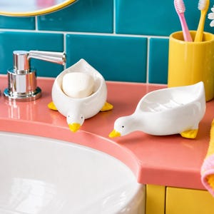May include: Two white ceramic duck-shaped soap dishes with yellow feet and beaks are on a pink countertop. One dish holds a bar of soap. A yellow toothbrush holder and a chrome faucet are also visible.