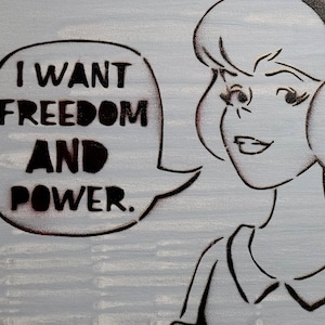 May include: A light blue painting features a woman's face and a speech bubble. The speech bubble contains the words "I WANT FREEDOM AND POWER." The artwork is done in a stencil style with black outlines.