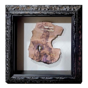 May include: A framed artwork featuring a black and white drawing of a woman in a vintage dress holding a fan. The drawing is on a piece of paper that has been torn and aged, giving it a distressed look. The frame is black with ornate detailing.