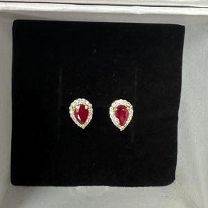 Può includere: Orecchini a lobo a forma di goccia, con una gemma rossa vibrante circondata da piccole pietre trasparenti. Gli orecchini sono incastonati in metallo color oro e presentati in un cofanetto bianco con interno in velluto nero. Un gioiello.