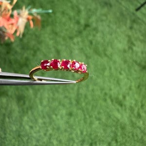 Op de afbeelding: Een gouden ring met vijf ovaal geslepen, helderrode edelstenen. De ring wordt door een pincet tegen een groene grasachtergrond gehouden. De edelstenen zijn gelijkmatig verdeeld in een delicate gouden band.