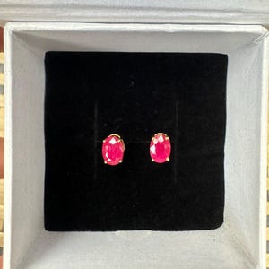 Pode incluir: Um par de brincos de rubi ovais em ouro, apresentados numa caixa de joias branca com interior de veludo preto. Os brincos são de cor vermelha vibrante.