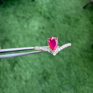 Pu&ograve; includere: Un anello d'oro con una gemma rossa a forma di pera. La fascia dell'anello &egrave; ornata da piccole gemme bianche scintillanti, che formano un motivo a forma di V. L'anello &egrave; tenuto da una pinzetta su uno sfondo verde.