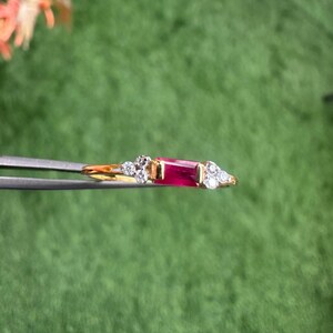 May include: A gold ring with a central rectangular ruby and smaller, clear gemstones. The ring is held by tweezers against a green backdrop, highlighting the contrast of the red and clear stones and the gold setting.