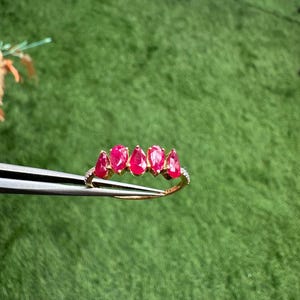 Pode incluir: Um anel de ouro com cinco rubis em forma de p&ecirc;ra, dispostos em linha. O anel &eacute; adornado com pequenas pedras preciosas transparentes ao longo da banda, criando um efeito cintilante. O anel &eacute; segurado por uma pin&ccedil;a.