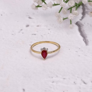 Puede incluir: Un anillo de oro con una piedra preciosa roja oscura en forma de pera, acentuada por pequeñas piedras claras. El anillo está sobre un fondo blanco con delicadas flores blancas en la esquina superior derecha.