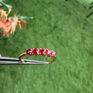 Op de afbeelding: Een gouden ring met vijf ovaal geslepen, helderrode edelstenen. De ring wordt door een pincet tegen een groene achtergrond gehouden. De edelstenen zijn gelijkmatig over de band verdeeld.