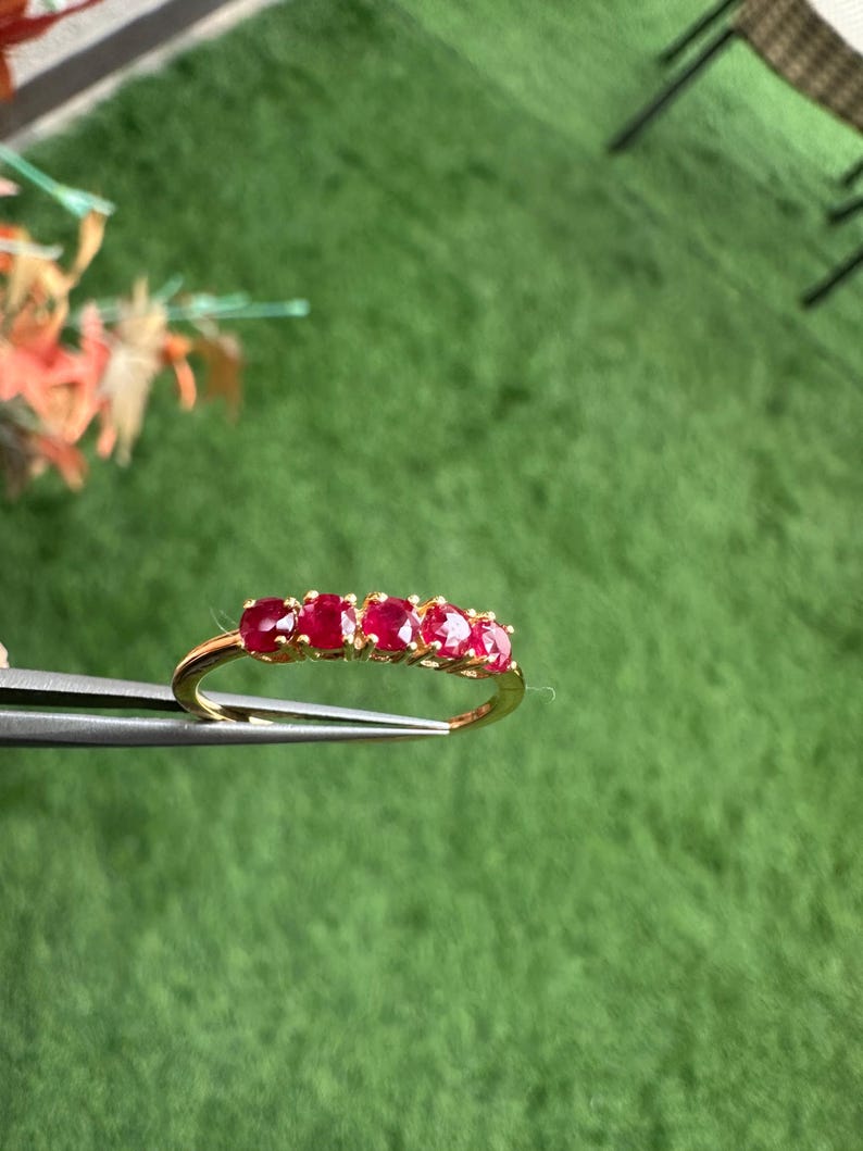 Op de afbeelding: Een gouden ring met vijf ovaal geslepen, helderrode edelstenen. De ring wordt door een pincet tegen een groene achtergrond gehouden. De edelstenen zijn gelijkmatig over de band verdeeld.