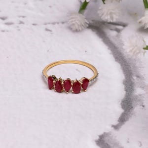 Pode incluir: Um anel de ouro com seis pedras preciosas vermelhas em forma de gota. A fina banda de ouro tem pequenas pedras transparentes na borda superior. O fundo &eacute; uma superf&iacute;cie marmoreada com flores brancas.