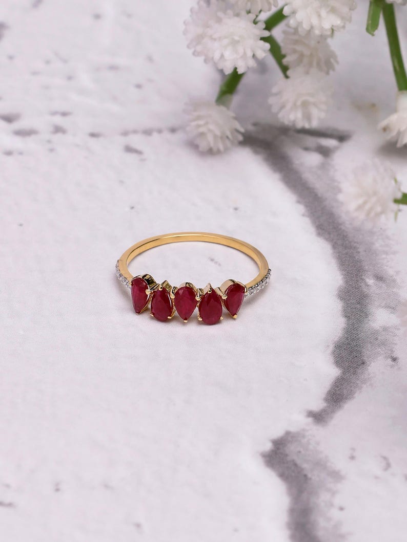 Pode incluir: Um anel de ouro com seis pedras preciosas vermelhas em forma de gota. A fina banda de ouro tem pequenas pedras transparentes na borda superior. O fundo &eacute; uma superf&iacute;cie marmoreada com flores brancas.