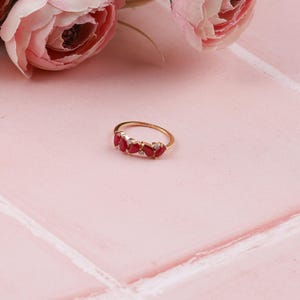 May include: A gold ring with a row of five red teardrop-shaped gemstones and small clear stones. The ring is set against a pink background with pink flowers in the background.
