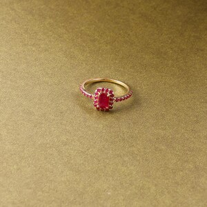 以下が含まれることがあります： 長方形の赤い宝石を中央に配し、周囲を小さな赤い石で囲んだゴールドリング。リングのバンドにも小さな赤い石が飾られています。リングは金色の背景に配置されています。