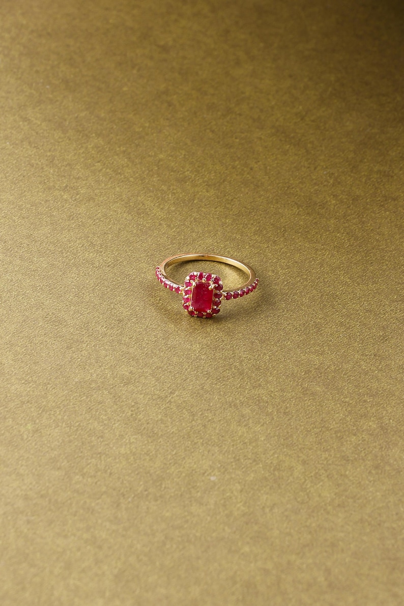 以下が含まれることがあります： 長方形の赤い宝石を中央に配し、周囲を小さな赤い石で囲んだゴールドリング。リングのバンドにも小さな赤い石が飾られています。リングは金色の背景に配置されています。