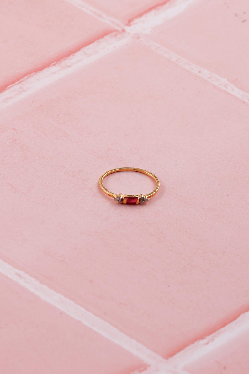 May include: A gold ring featuring a rectangular red gemstone, with two smaller clear stones either side. The ring is set on a pink tiled surface. The design is simple and elegant.