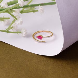 May include: A gold ring featuring a pear-shaped, red gemstone. The ring's band is set with small, clear stones. The ring is presented on a white surface, with a sprig of white flowers in the background.