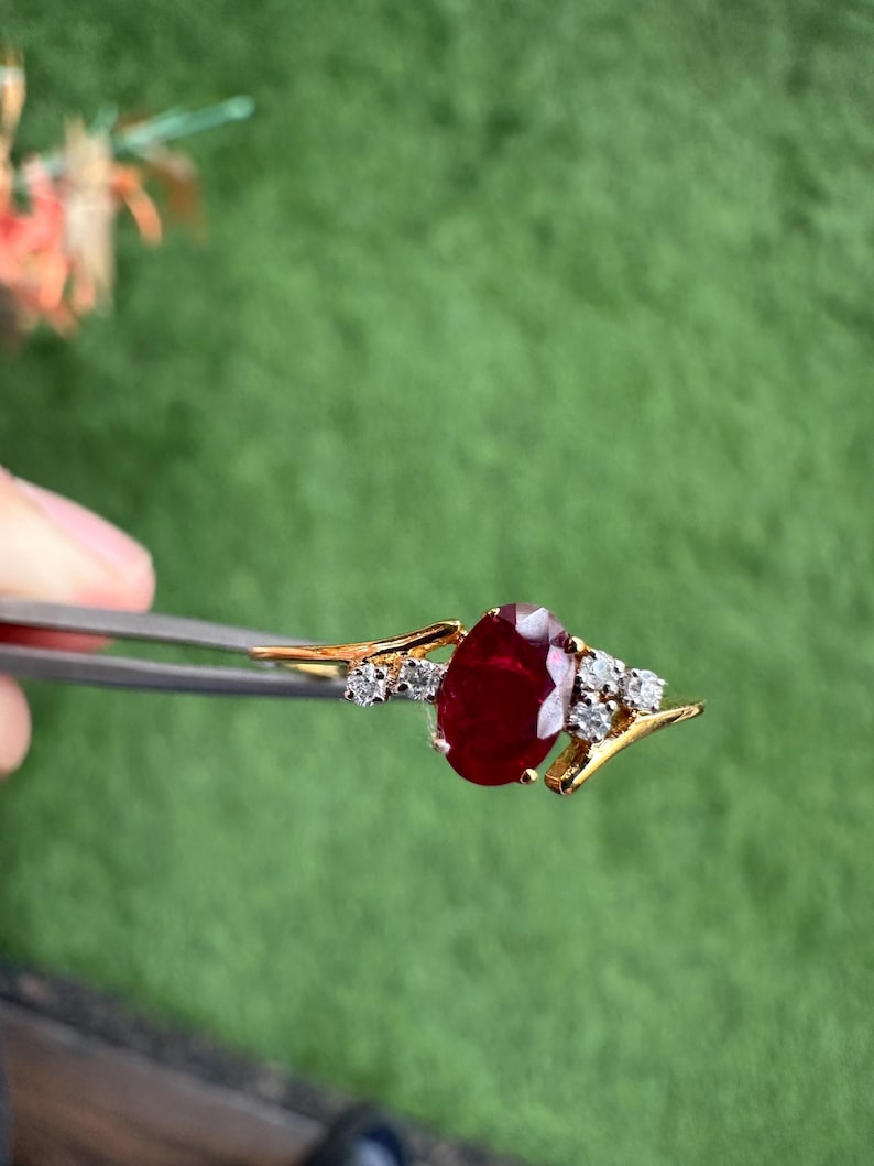 May include: A gold ring with a large, oval-cut ruby gemstone. The ruby is a deep red colour and is accented by smaller, clear gemstones. The ring is held by tweezers against a green background.
