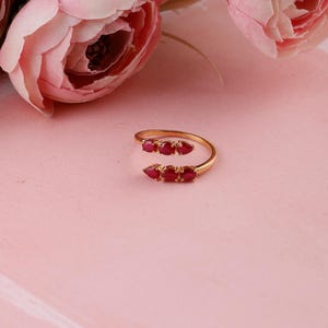 May include: A gold-colored ring with five red gemstones. The ring has an open design, with the gemstones arranged in two clusters. The ring is set against a pink background, with pink flowers in the background.
