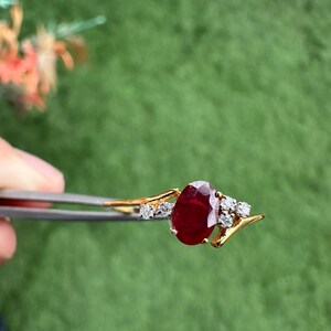 May include: A gold ring with a large, oval-cut ruby gemstone. The ruby is a deep red colour and is surrounded by smaller, clear diamonds. The ring is held by tweezers against a green background.
