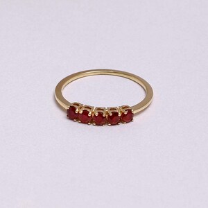 Op de afbeelding: Een gouden ring met vijf ovale, helderrode edelstenen. De edelstenen zijn in een rij geplaatst over de bovenkant van de ring. Het ontwerp is eenvoudig en elegant, waardoor de edelstenen opvallen.