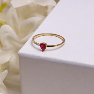 Op de afbeelding: Een gouden ring met een peervormige, rode edelsteen en kleine, heldere accentstenen. De ring ligt op een wit oppervlak, met zachte, vage witte bloemen op de achtergrond. De ringband is dun en delicaat.