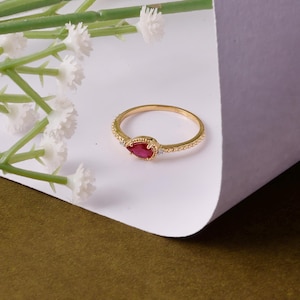 Peut inclure: Une bague en or avec une pierre précieuse rouge en forme de poire et de petites pierres blanches. L'anneau de la bague a un motif texturé. Des fleurs blanches et des tiges vertes sont en arrière-plan, avec un fond blanc et vert olive.