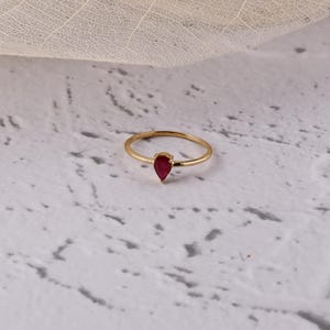 May include: A delicate gold ring featuring a teardrop-shaped, deep red gemstone. The ring's band is thin and simple, with the gemstone set at the center. The ring is displayed on a textured, light-colored surface.