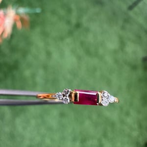 May include: A gold ring with a rectangular, deep red gemstone at its centre, complemented by clusters of small, round, clear gemstones. The ring is set against a blurred green backdrop.