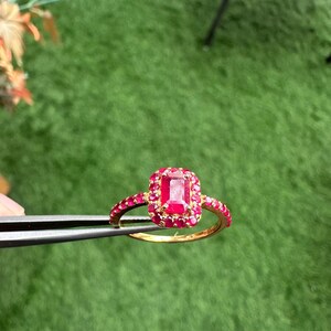 以下が含まれることがあります： 大きな長方形のルビー色の宝石を、小さな宝石で囲んだゴールドリング。リングのバンドにもルビー色の石が飾られています。リングは緑色の背景に対してピンセットで保持されています。