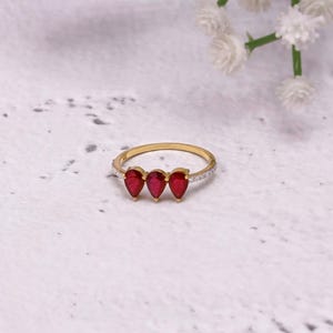Könnte beinhalten: Ein goldener Ring mit drei tropfenförmigen, tiefroten Edelsteinen. Das dünne Goldband hat kleine, klare Steine, die auf der oberen Hälfte eingesetzt sind. Der Ring ist vor einem weiß-grau gesprenkelten Hintergrund platziert.