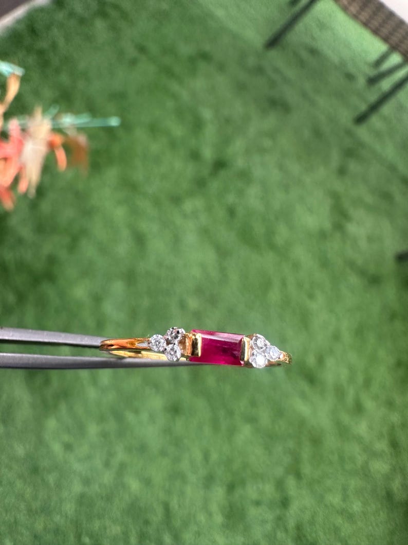 May include: A gold ring with a central rectangular ruby and surrounding clear gemstones. The ring is held by tweezers. The background is green, highlighting the ring's design and the sparkle of the gemstones.