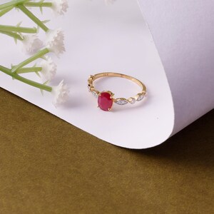 Op de afbeelding: Een gouden ring met een ovale, rode edelsteen. De ring heeft een fijne band met kleine diamantaccenten. De ring is geplaatst tegen een witte en olijfgroene achtergrond. Witte bloemen zijn op de achtergrond.