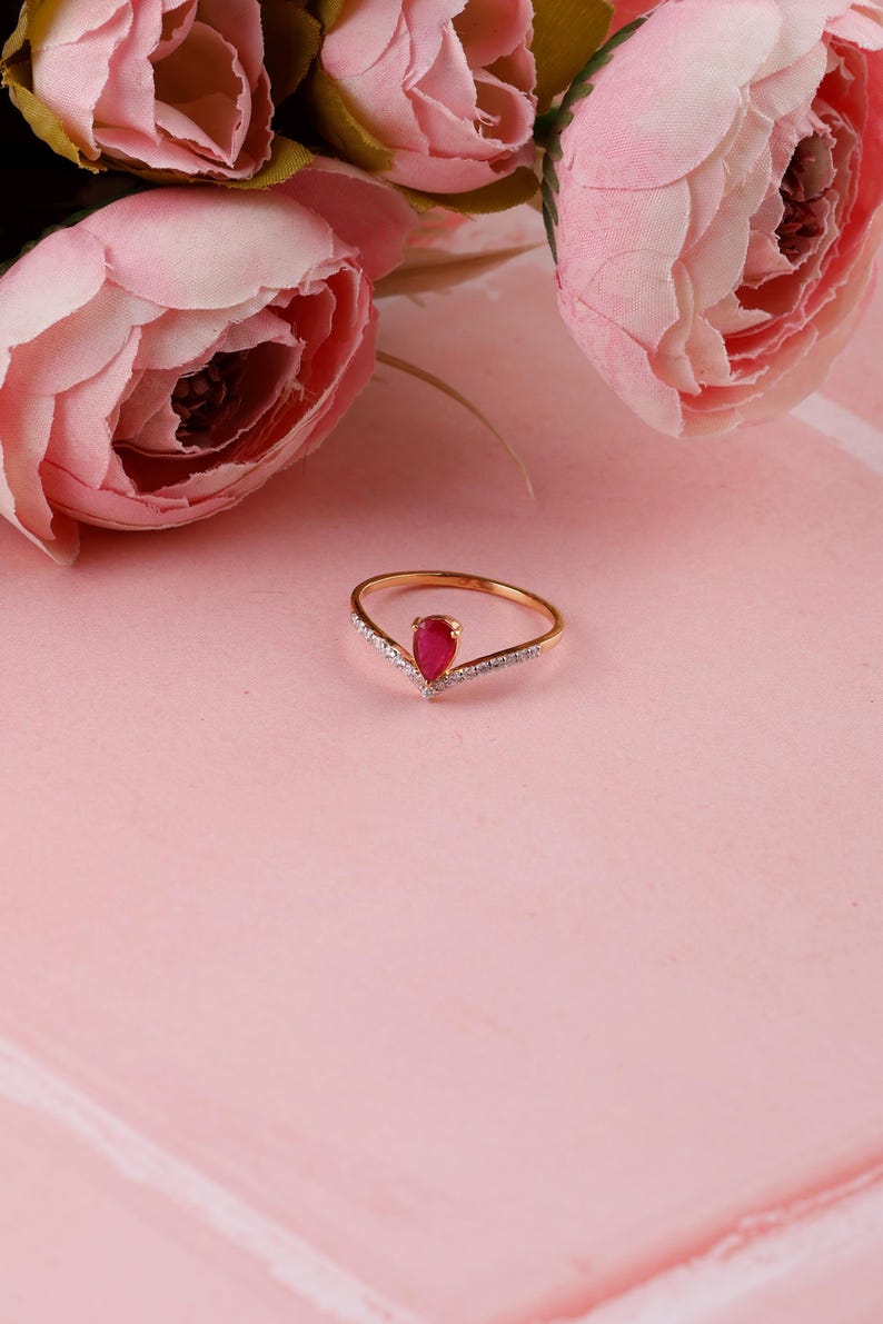 Pu&ograve; includere: Un anello d'oro con una gemma rossa a forma di goccia al centro. L'anello ha un design a forma di V con piccole pietre trasparenti lungo i bordi. Fiori rosa sullo sfondo, su una superficie rosa.