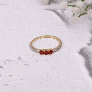 May include: A gold ring featuring three small, red gemstones at the centre. The thin band is embellished with small, clear stones. The ring is presented on a white, textured surface with white flowers in the background.