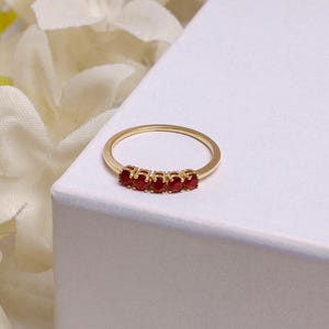Op de afbeelding: Een goudkleurige ring met vijf kleine, ronde, rode edelstenen in een rij. De ring wordt gepresenteerd op een wit oppervlak, met vage witte bloemen op de achtergrond.