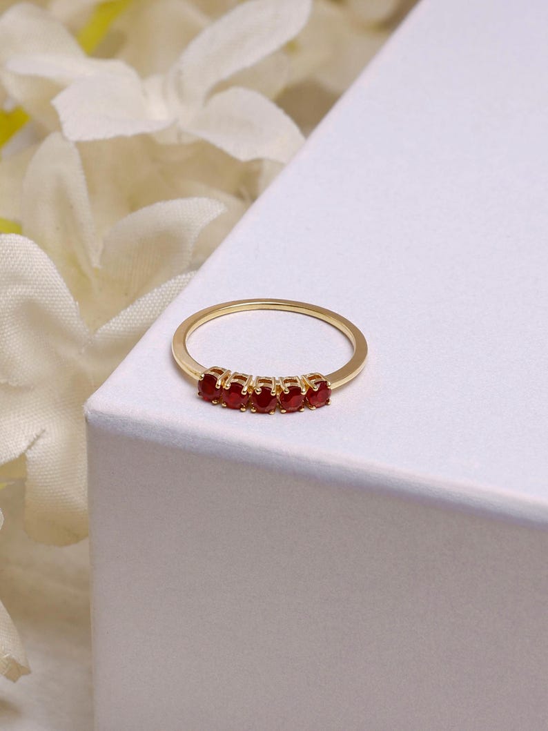 Op de afbeelding: Een goudkleurige ring met vijf kleine, ronde, rode edelstenen in een rij. De ring wordt gepresenteerd op een wit oppervlak, met vage witte bloemen op de achtergrond.