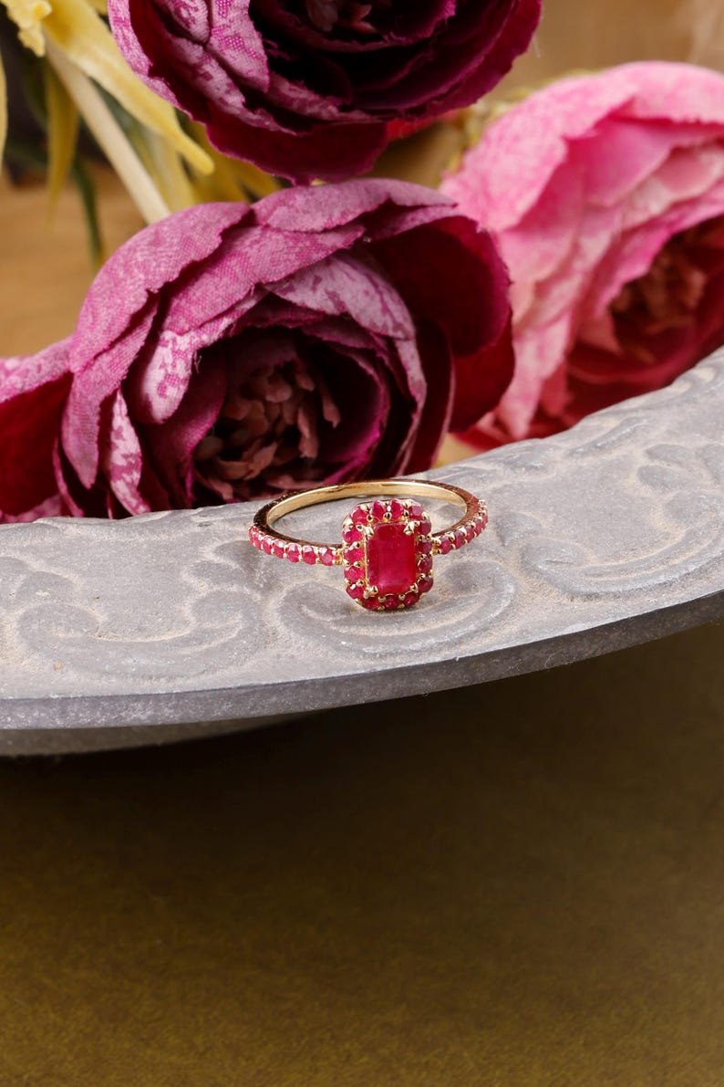 以下が含まれることがあります： 長方形のルビー色の中心石と、小さな赤い石で囲まれた金の指輪。指輪は、グレーの質感のある表面に置かれ、背景にはピンクと紫の花があります。