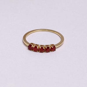 Op de afbeelding: Een gouden ring met vijf ovale, helderrode edelstenen. De edelstenen zijn in een rij geplaatst over de bovenkant van de ring. Het ontwerp is eenvoudig en elegant, waardoor de edelstenen opvallen.