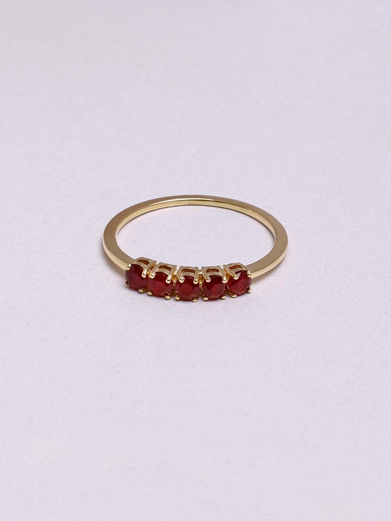 Op de afbeelding: Een gouden ring met vijf ovale, helderrode edelstenen. De edelstenen zijn in een rij geplaatst over de bovenkant van de ring. Het ontwerp is eenvoudig en elegant, waardoor de edelstenen opvallen.