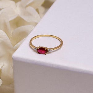 Op de afbeelding: Een gouden ring met een ovale, rode robijn in het midden en twee kleinere, heldere edelstenen. De ring heeft een dunne, gepolijste band. De ring is geplaatst op een witte achtergrond.