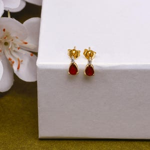 Puede incluir: Un par de pendientes dorados con una gema roja oscura en forma de lágrima y una pequeña piedra transparente encima. Los pendientes se muestran sobre una superficie blanca, con una flor blanca en el fondo.