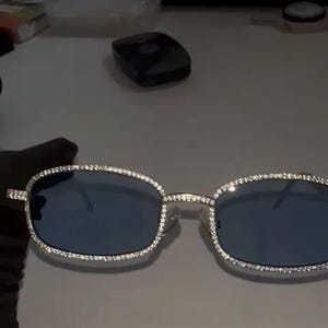 May include: Silver-framed sunglasses with blue lenses. The frames are encrusted with small, sparkling crystals around the edges. The rectangular shape and crystal embellishments give the sunglasses a luxurious appearance.
