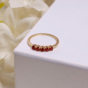May include: A gold-colored ring featuring five small, round, red gemstones set in a row. The ring is displayed on a white surface, with soft, blurred white flowers in the background.