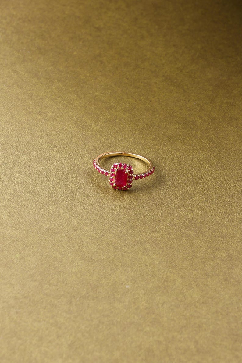 以下が含まれることがあります： 長方形のルビー色の中心石が付いたゴールドリング。バンドにも小さなルビー色の石が飾られています。リングは金色の背景にセットされています。