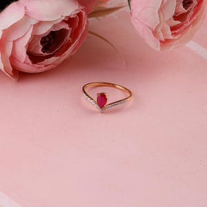 Pu&ograve; includere: Un anello d'oro con una gemma rossa a forma di goccia al centro. L'anello ha un design a forma di V con piccole pietre trasparenti lungo i bordi. Fiori rosa sullo sfondo, su una superficie rosa.