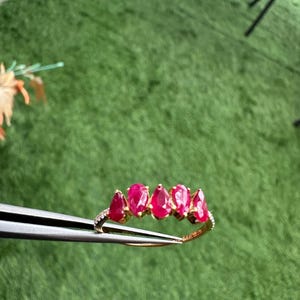 Pode incluir: Um anel de ouro com cinco pedras preciosas vermelhas vibrantes em forma de p&ecirc;ra. O anel &eacute; adornado com pequenas pedras transparentes ao longo da banda. O anel &eacute; segurado por uma pin&ccedil;a sobre um fundo verde.