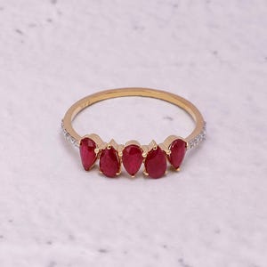 Pode incluir: Um anel de ouro com cinco pedras preciosas vermelhas em forma de gota. A banda fina &eacute; adornada com pequenas pedras transparentes em ambos os lados das pedras vermelhas. O anel &eacute; apresentado sobre um fundo branco e cinzento.