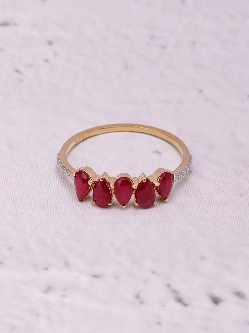 Pode incluir: Um anel de ouro com cinco pedras preciosas vermelhas em forma de gota. A banda fina &eacute; adornada com pequenas pedras transparentes em ambos os lados das pedras vermelhas. O anel &eacute; apresentado sobre um fundo branco e cinzento.