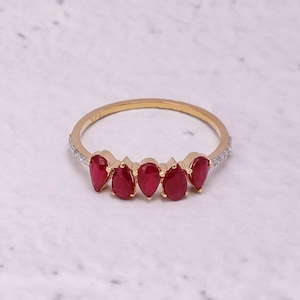 Pode incluir: Um anel de ouro com cinco pedras preciosas vermelhas em forma de gota. A banda &eacute; adornada com pequenas pedras transparentes em ambos os lados das pedras vermelhas. O anel est&aacute; sobre um fundo branco e cinza.