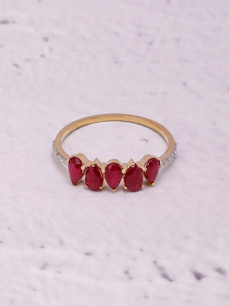 Pode incluir: Um anel de ouro com cinco pedras preciosas vermelhas em forma de gota. A banda &eacute; adornada com pequenas pedras transparentes em ambos os lados das pedras vermelhas. O anel est&aacute; sobre um fundo branco e cinza.